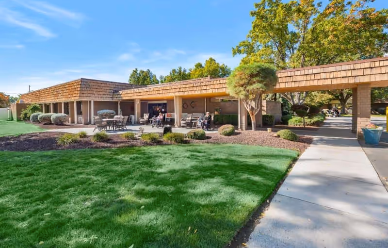 Outdoor courtyard and covered walkway of a retirement community with patio seating, manicured lawn, shrubs and a sidewalk.