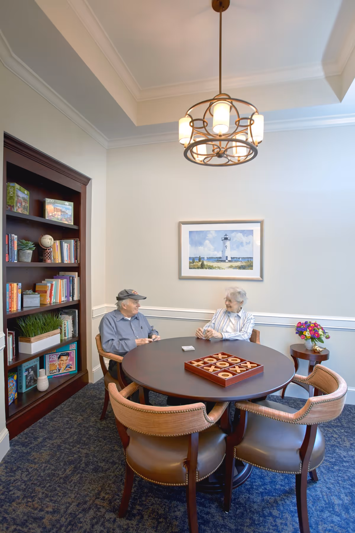 Two senior residents seated at a round table playing tic-tac-toe in a well-lit common room with a bookshelf, framed lighthouse painting, and overhead chandelier.
