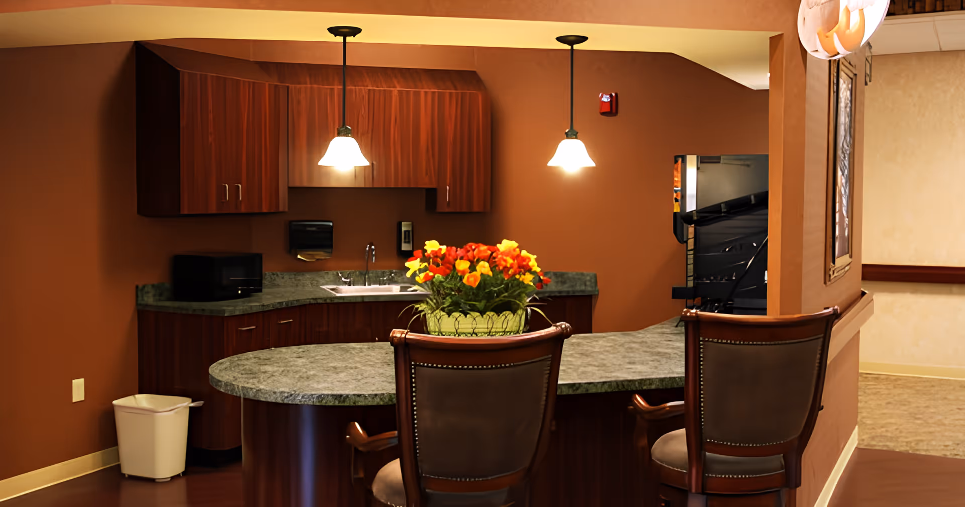 Interior view of a kitchen area with dark wood cabinets, a green marble countertop, two pendant lights hanging from the ceiling, a microwave, a sink, and a vase with red and yellow flowers on the counter. Two wooden chairs with cushioned seats are positioned at the counter. A trash bin is placed on the floor to the left.