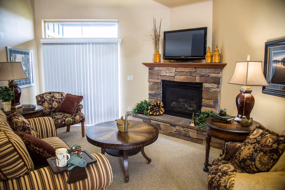 A cozy living room with a stone fireplace topped by a flat-screen TV. The room features a round wooden coffee table, a striped sofa with cushions, two patterned armchairs, side tables with lamps, and framed artwork on the walls. Vertical blinds cover a window letting in natural light.