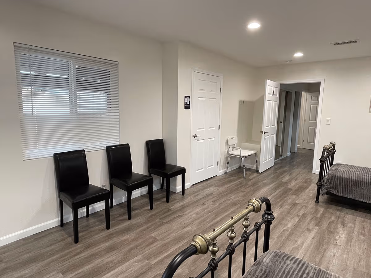 Interior view of a senior living facility room with three black chairs lined up against a wall with a window covered by blinds. There are two beds with metal frames and gray bedding visible, a white chair near a closed door, and a hallway leading to other rooms. The floor is wood-style laminate and the walls are painted white.
