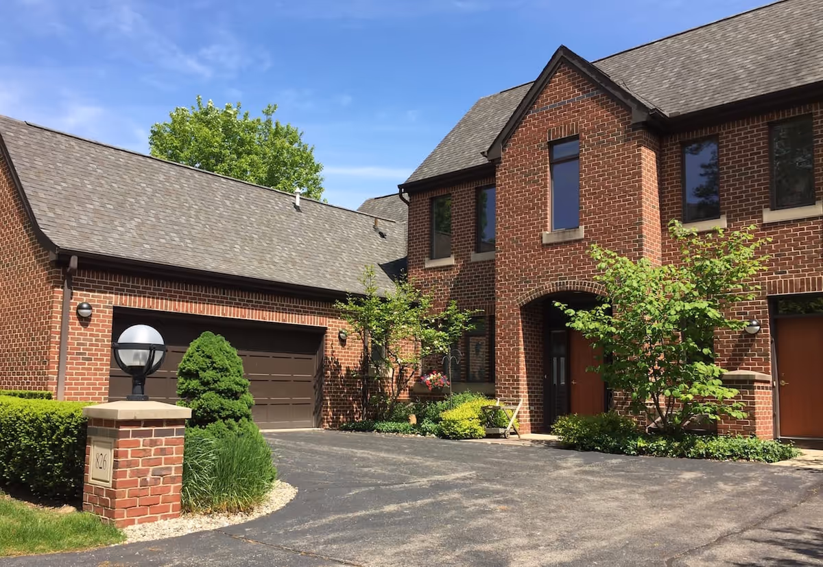 Exterior view of a brick building with a gray shingled roof, featuring a garage with a dark brown door, several windows, and a main entrance under an archway. The driveway is paved and bordered by green shrubs, small trees, and a brick pillar with a round lamp on top. The sky is clear and blue.