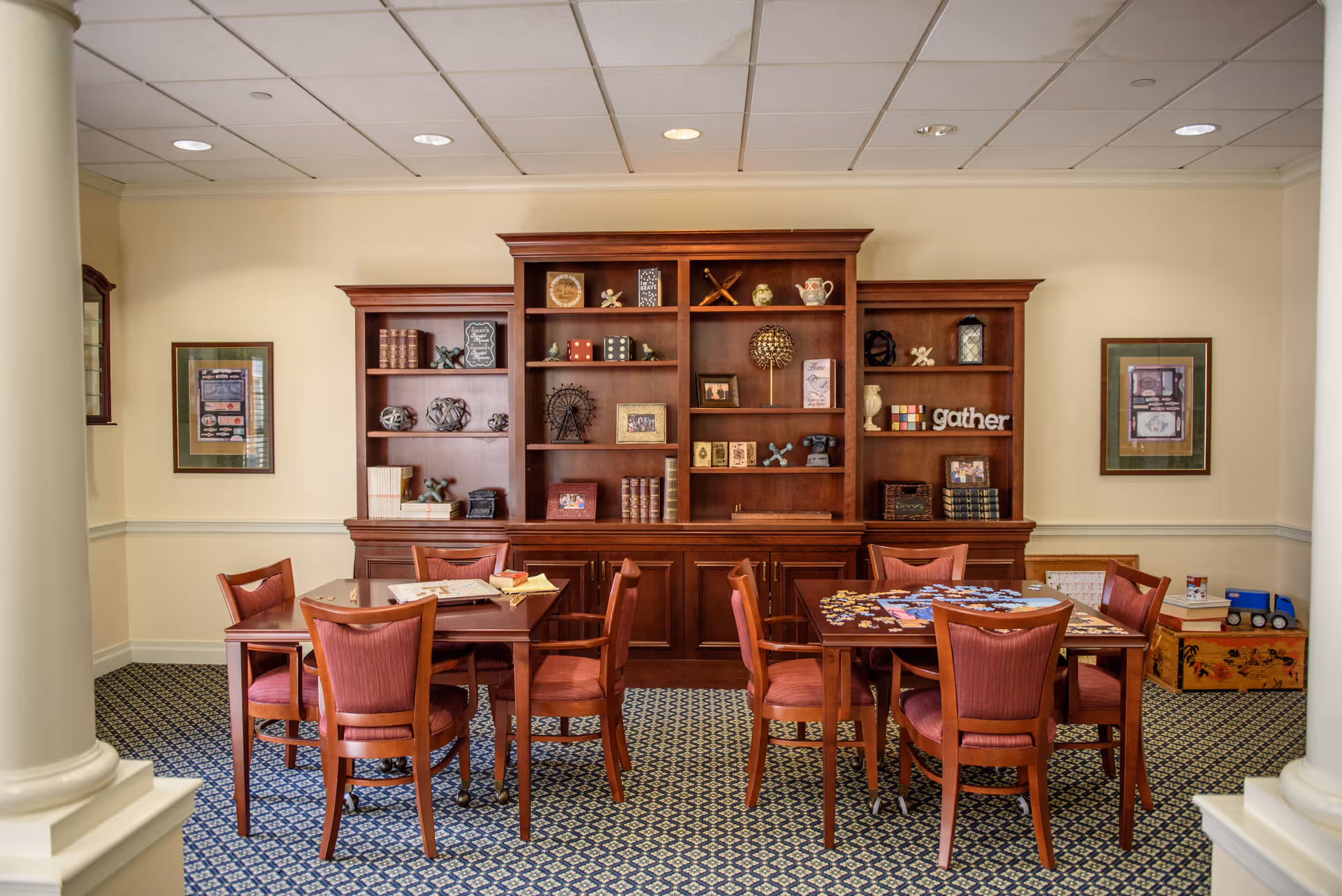 A cozy interior room with two wooden tables surrounded by wooden chairs with red cushions. One table has a partially completed jigsaw puzzle, and the other has a board game. Behind the tables is a large wooden bookshelf filled with decorative items, books, and framed pictures. The room has patterned carpet flooring, cream-colored walls, and framed artwork on the walls.