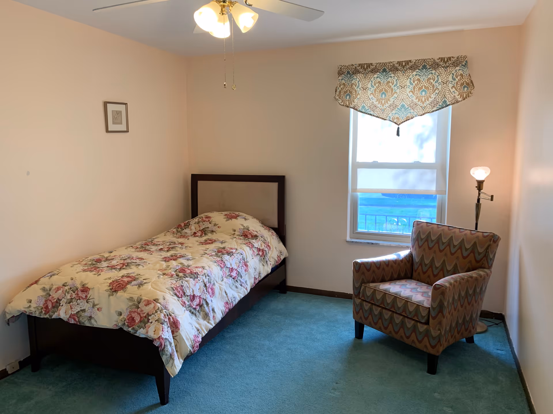 A small bedroom with a single bed covered with a floral comforter, a patterned armchair, a floor lamp, a window with a decorative valance, and a ceiling fan with lights.