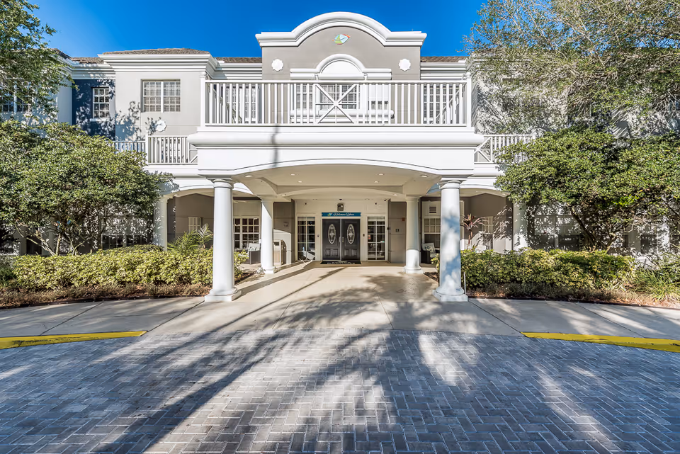 Front entrance of a two-story white senior living building with columns, a covered driveway, balcony, and landscaped shrubs.