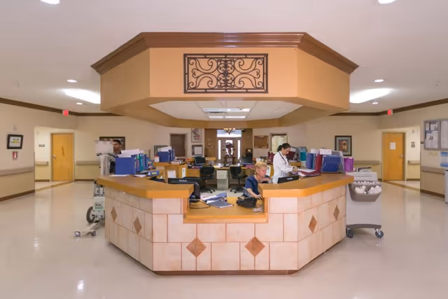 A wide view of a healthcare facility's central nurse station with two staff members working. The station has a tiled front with a wooden countertop and is surrounded by a clean, well-lit hallway with doors and framed artwork on the walls.