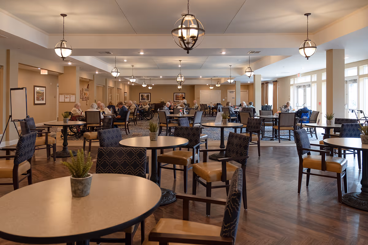 A spacious dining room in a senior living facility with round tables and chairs arranged neatly. Several elderly residents are seated at tables in the background, some in wheelchairs, engaged in conversation or dining. The room is well-lit with hanging pendant lights and large windows allowing natural light to enter. Small potted plants decorate the tables.