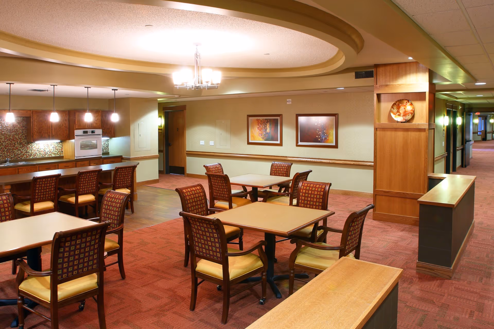 A well-lit dining area in a senior living facility with several square tables and cushioned chairs arranged neatly. The room features a kitchen area with wooden cabinets, a built-in oven, and pendant lights hanging above a counter with bar stools. The walls are decorated with framed abstract artwork, and the ceiling has a recessed circular design with a chandelier. The flooring is a combination of carpet and wood, and a hallway is visible in the background.