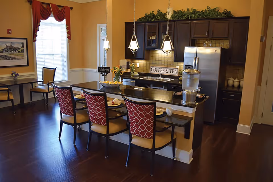 A kitchen and dining area in a senior living facility with a dark wood floor, a kitchen island with four chairs featuring red patterned backs, and a countertop set with plates and napkins. The kitchen has dark wood cabinets, a stainless steel refrigerator, a water dispenser on the counter, and a sign that reads 'COFFEE & TEA'. There is a window with red curtains and a small table with two chairs in the background.