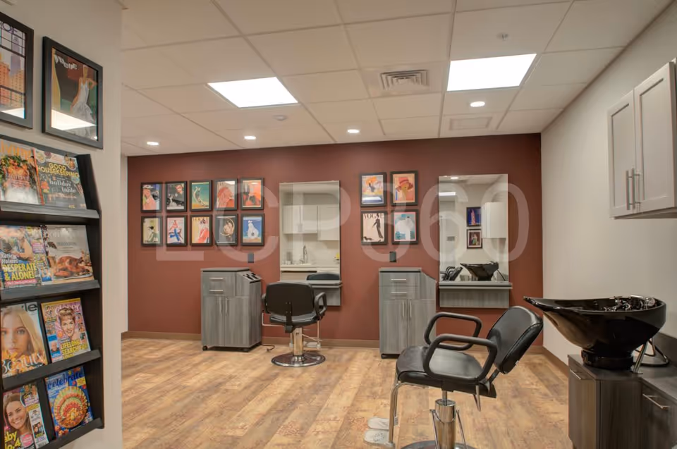 Interior of a salon area in Mercer Hill at Doylestown featuring two black salon chairs, two mirrors mounted on a maroon wall, small cabinets beneath each mirror, a black hair washing sink on the right, and a magazine rack filled with various magazines on the left. The floor is wooden and the ceiling has recessed lighting.