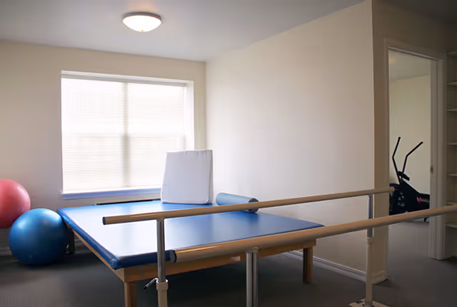 A rehabilitation/therapy room with parallel walking bars over a padded blue therapy table, exercise balls, and an exercise bike visible in an adjoining room.