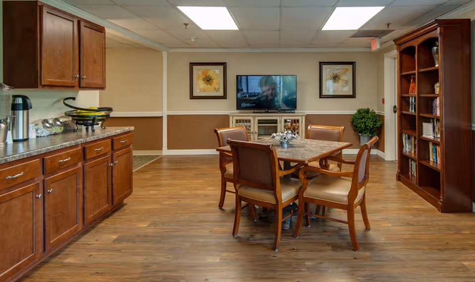 A cozy common area in Spencer Place featuring a wooden table with four chairs in the center, a flat-screen TV mounted on the wall with two framed floral pictures on either side, wooden cabinets and countertop on the left, and a tall wooden bookshelf filled with books and decorative items on the right. The room has wood flooring and a ceiling with fluorescent lighting.