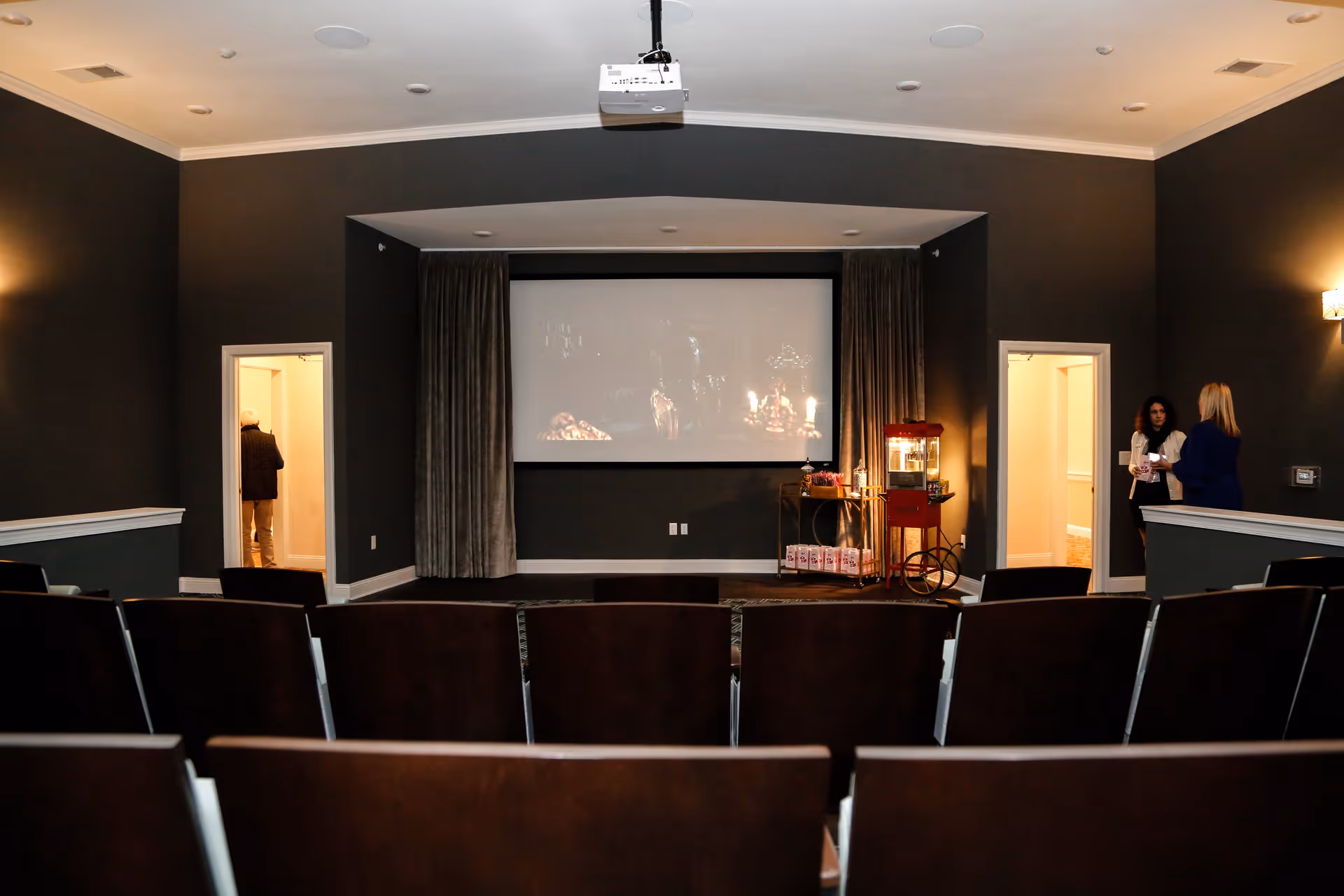 A small movie theater room with rows of dark wooden seats facing a large projection screen. There is a popcorn machine and a cart with snacks on the right side near the screen. Two women are standing and talking near a doorway on the right, and a person is visible through a doorway on the left. The walls are painted dark gray with white trim, and ceiling lights provide soft illumination.