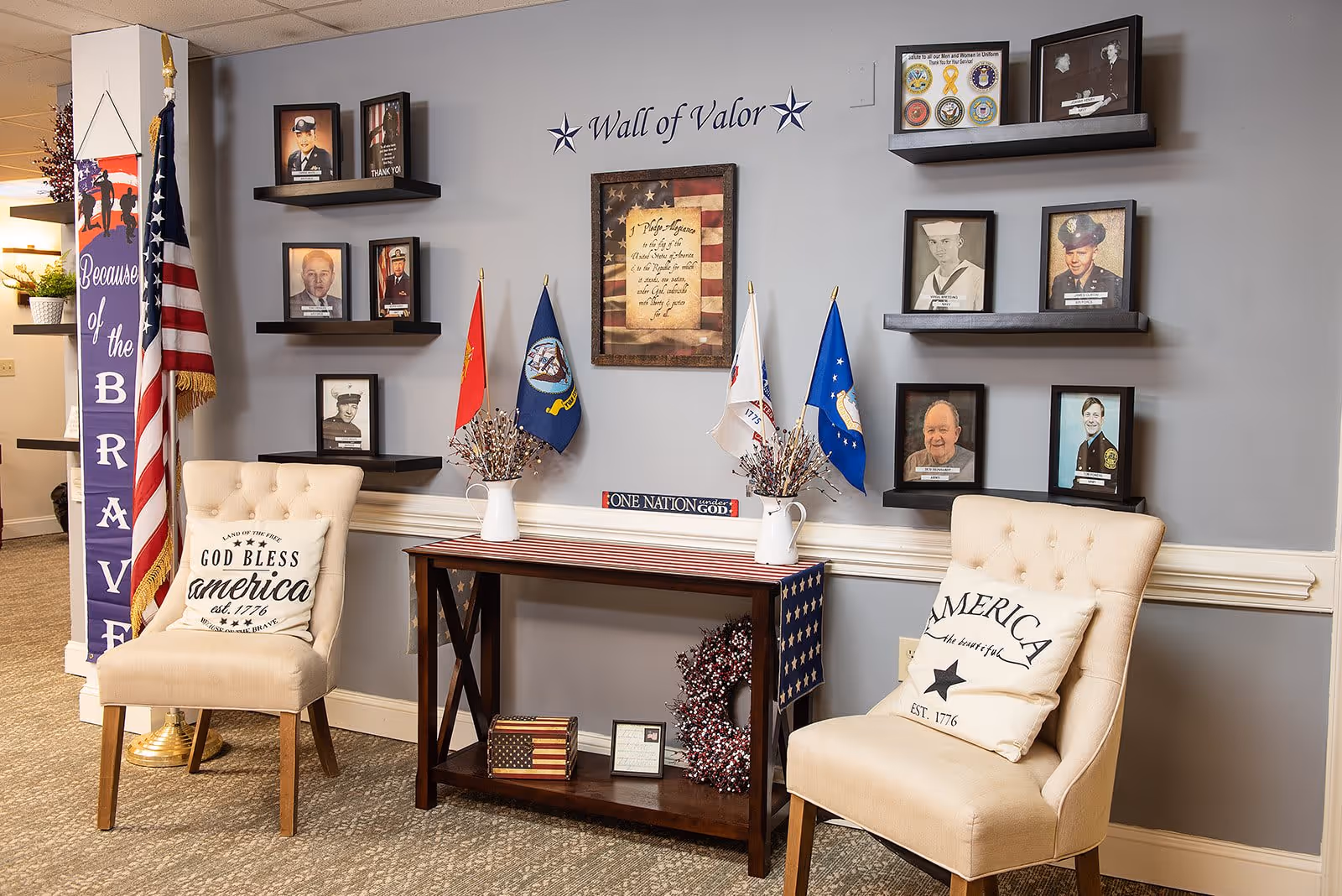 A patriotic-themed seating area in a senior living facility featuring two beige upholstered chairs with America-themed pillows. Between the chairs is a wooden table decorated with small American flags, a wreath, and framed items. The wall behind has a 'Wall of Valor' display with framed photos of veterans, military flags, and patriotic decor. An American flag stands to the left with a banner that reads 'Because of the Brave.'