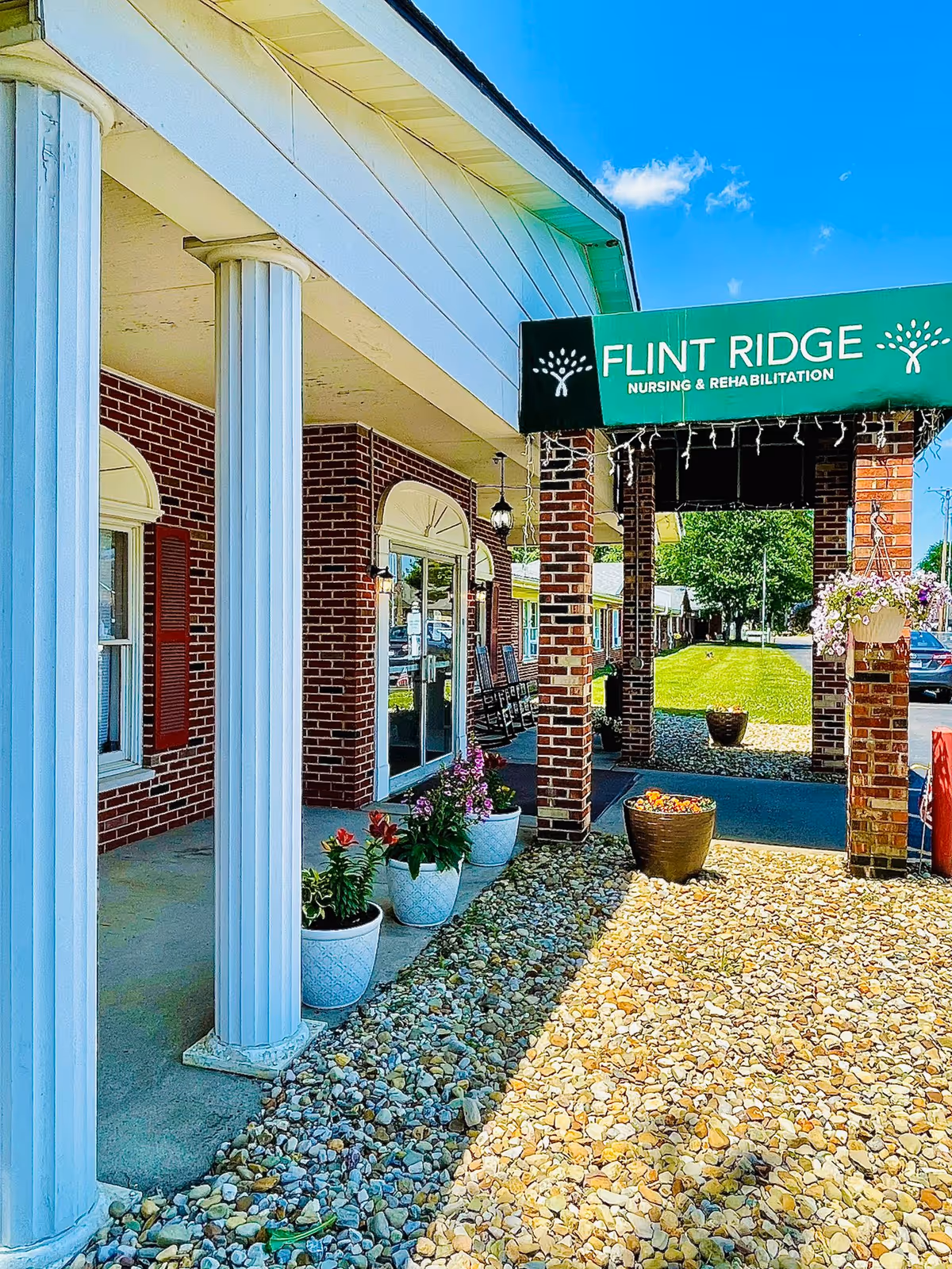 Exterior view of Flint Ridge Nursing and Rehabilitation facility showing a brick building with white columns, a green awning with the facility name, potted plants along the walkway, and a clear blue sky.
