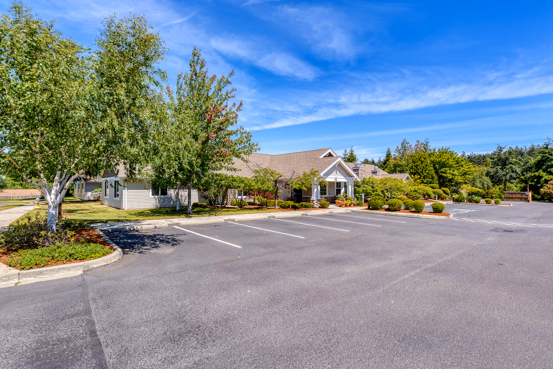 Exterior view of San Juan Villa Memory Care facility showing a single-story building with a pitched roof surrounded by trees and landscaped bushes. There is a large empty parking lot in front under a blue sky with some clouds.