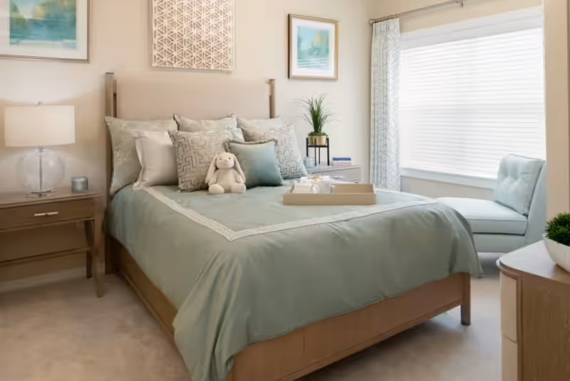 Cozy pastel bedroom with a bed, nightstands, plant, and window seating area.