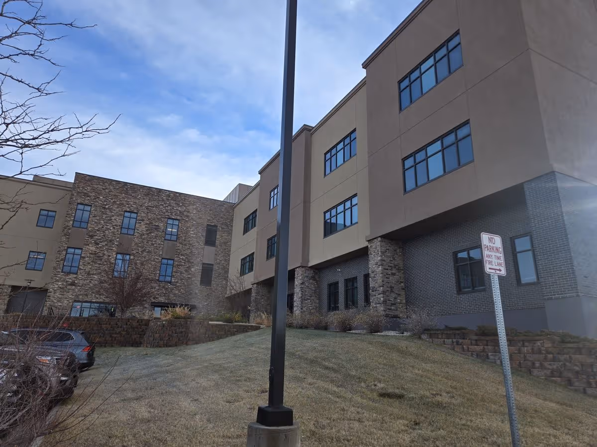 Exterior view of a multi-story senior living building with stone and stucco facade, windows, a landscaped slope, a lamppost, and a 'No Parking' sign.