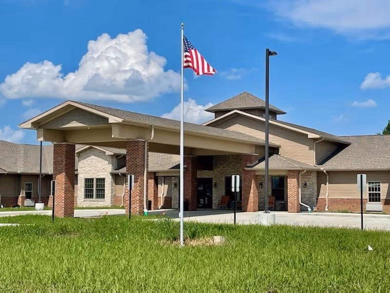 Exterior view of The Carriages of Rochester facility showing a single-story building with a covered entrance supported by brick columns, an American flag on a flagpole in front, and a bright blue sky with some clouds.