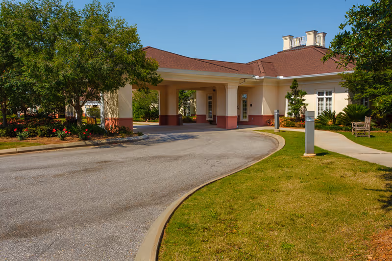 Exterior view of The Brennity at Daphne Assisted Living & Memory Care building entrance with a covered driveway, surrounded by green grass, trees, and shrubs under a clear blue sky.