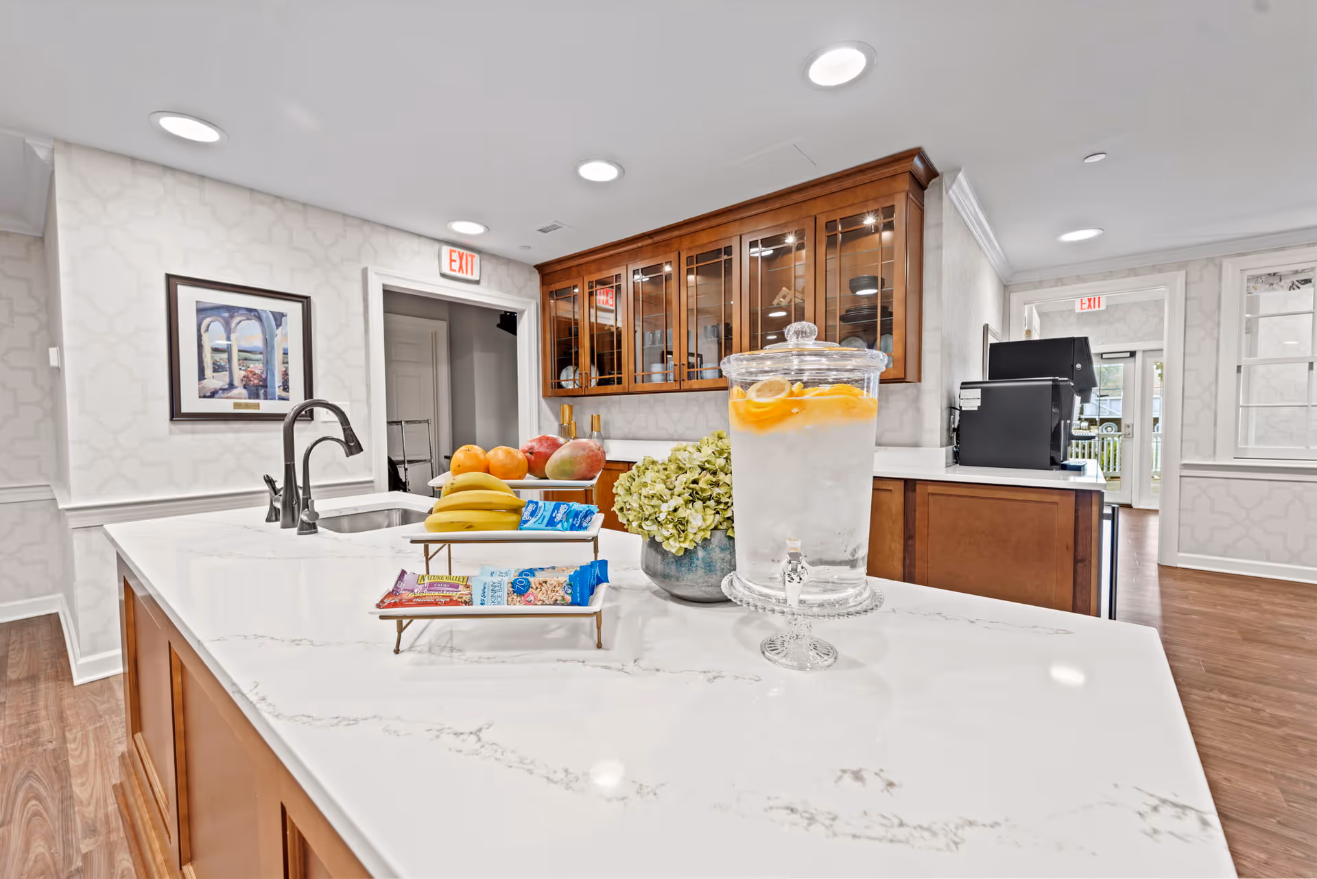 Bright kitchen area with a white marble countertop island featuring a glass dispenser filled with water and lemon slices, a bowl of bananas and other fruits, and a small tray with snack bars. The kitchen has wooden cabinets with glass doors, a sink with a modern faucet, and a coffee machine in the background. The walls are decorated with light patterned wallpaper and a framed picture. Two exit signs are visible above doorways.