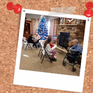Several seniors sit in folding chairs around a decorated Christmas tree and stone fireplace in a communal living room.
