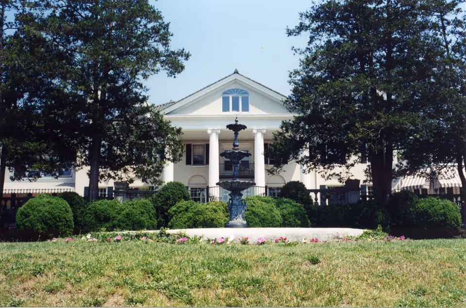 Front exterior view of a large white estate building with tall columns, a central fountain, surrounded by green bushes and trees under a clear blue sky.