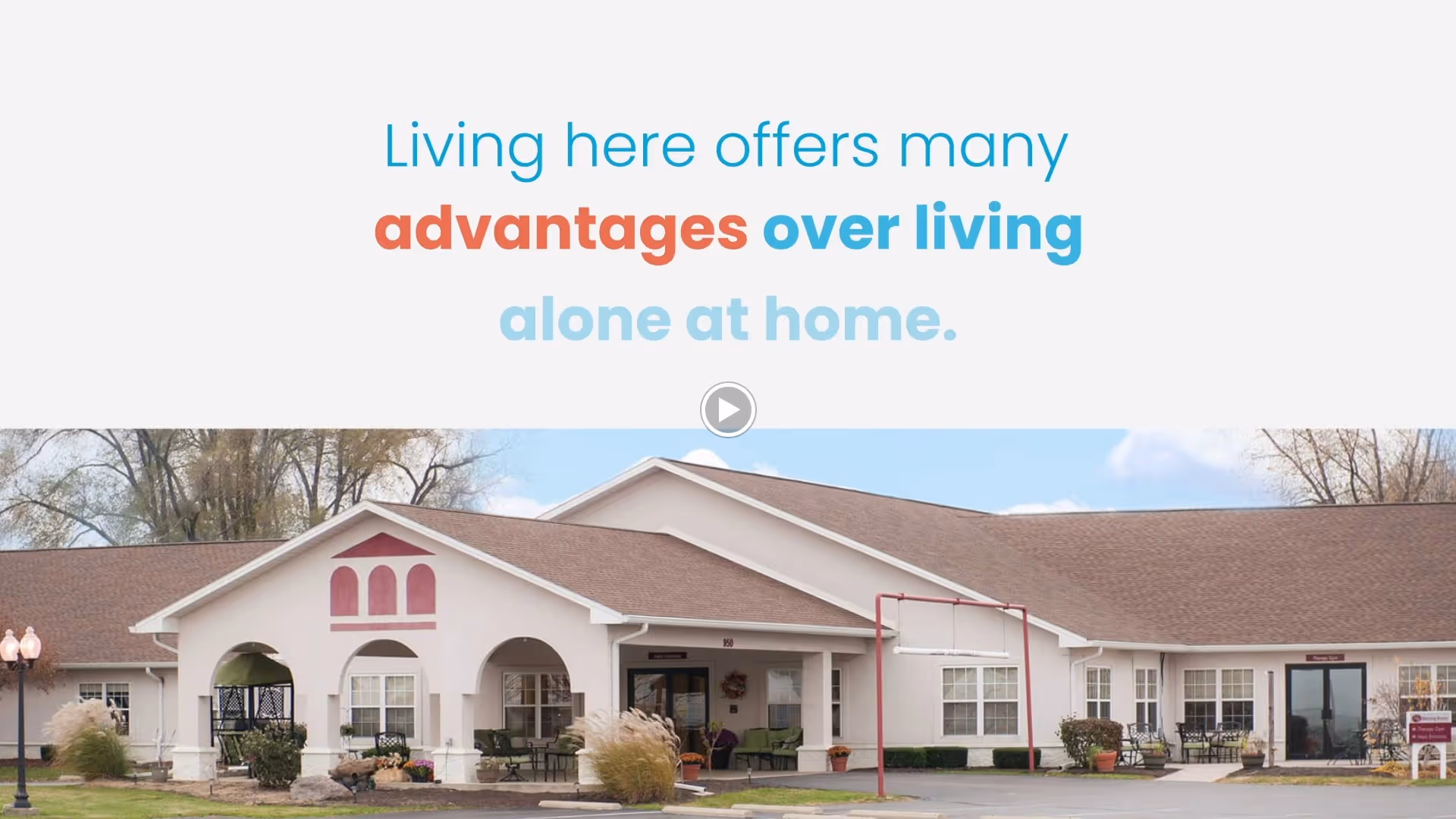 Front exterior of a single-story retirement community building with an arched entrance, outdoor seating, and a banner reading 'Living here offers many advantages over living alone at home.'