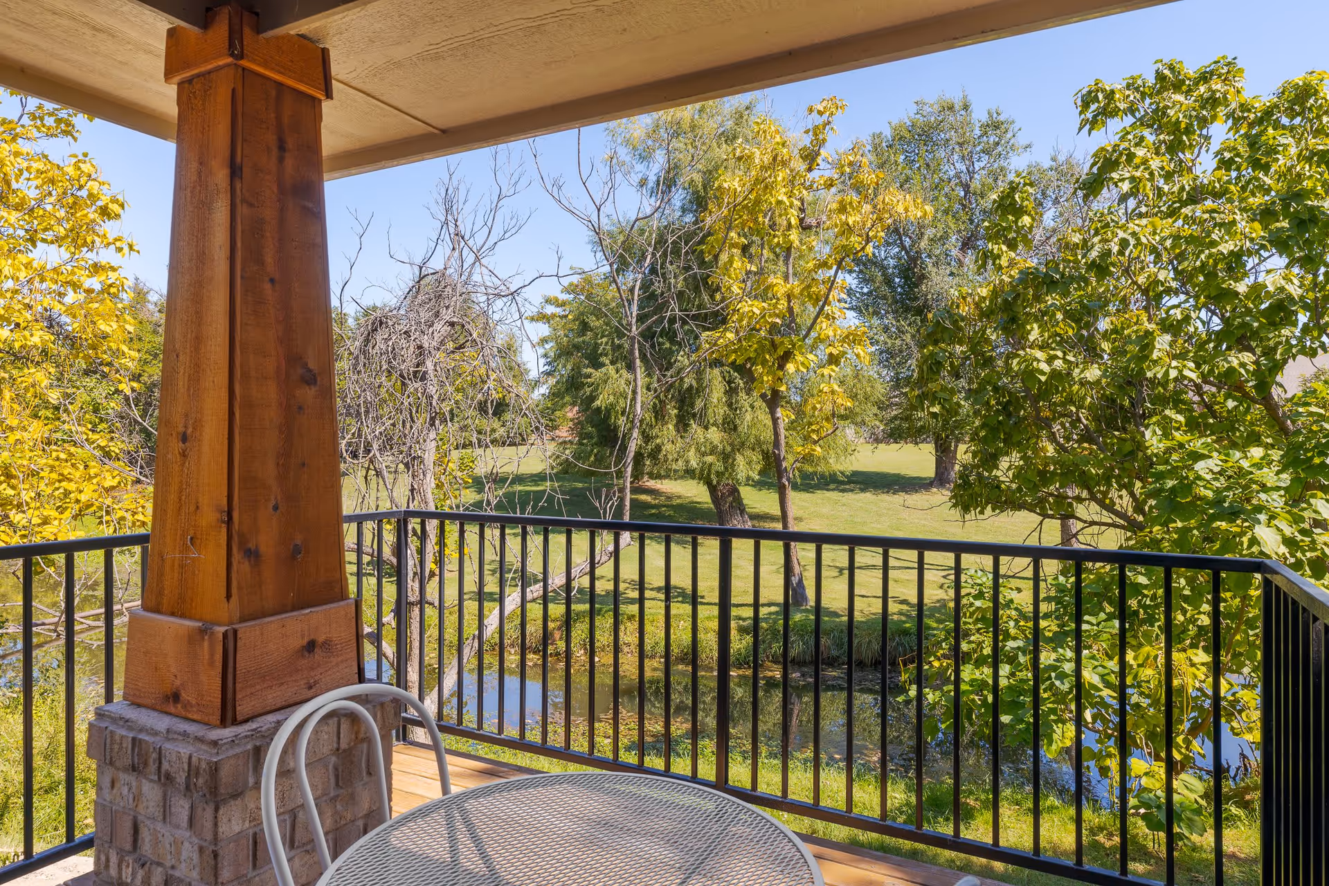View from a covered balcony with a metal table and chair, overlooking a grassy area with trees and a small pond under a clear blue sky.