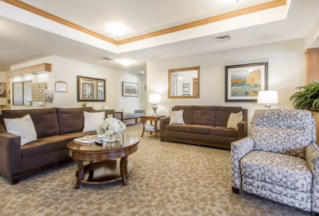 A well-lit senior living facility common area with two brown sofas, a patterned armchair, a round wooden coffee table with a flower arrangement and magazines, beige carpet, framed artwork on the walls, table lamps, and a large mirror.