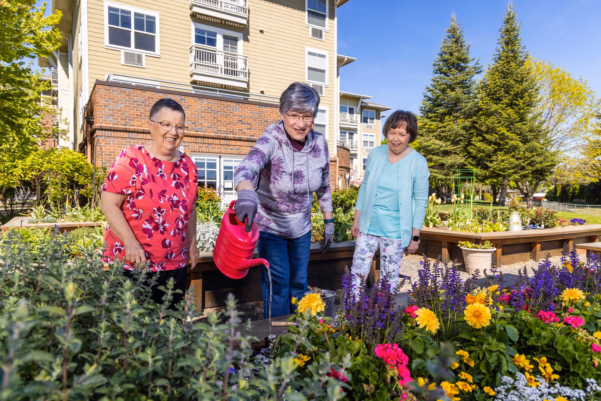 Three people tending a colorful raised flower garden outdoors in front of a multi-story residential building.