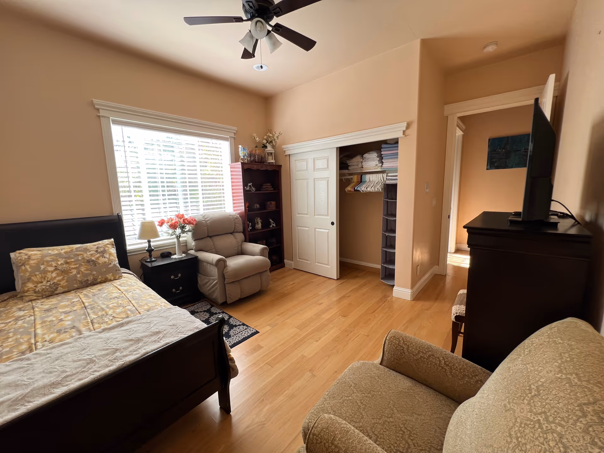 A cozy bedroom with a wooden bed featuring floral bedding, a beige recliner chair next to a window with white blinds, a small black nightstand with a lamp and flowers, a wooden bookshelf with decorative items, an open closet with neatly folded towels and hangers, a dark wooden dresser with a TV on top, and a beige upholstered armchair in the foreground. The room has light-colored walls and wooden flooring.