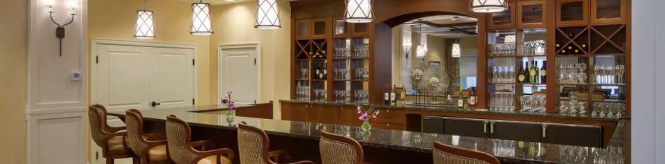 Elegant interior bar area with a long granite counter, bar stools, pendant lights, and glass-front cabinets stocked with glassware and bottles.