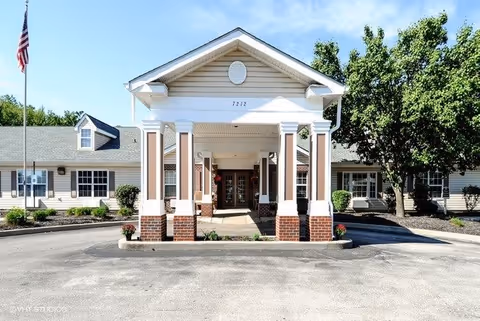Front exterior view of The Wellington at Southport building with a covered entrance supported by white columns, surrounded by trees and landscaping under a clear blue sky.