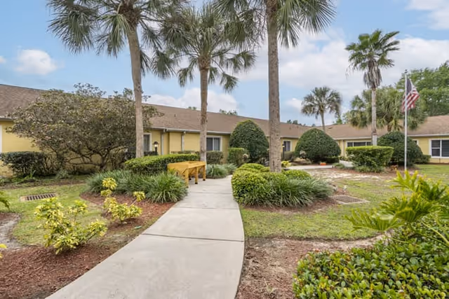 A paved walkway leading through a landscaped garden area with palm trees and various shrubs in front of a single-story yellow building under a partly cloudy sky.