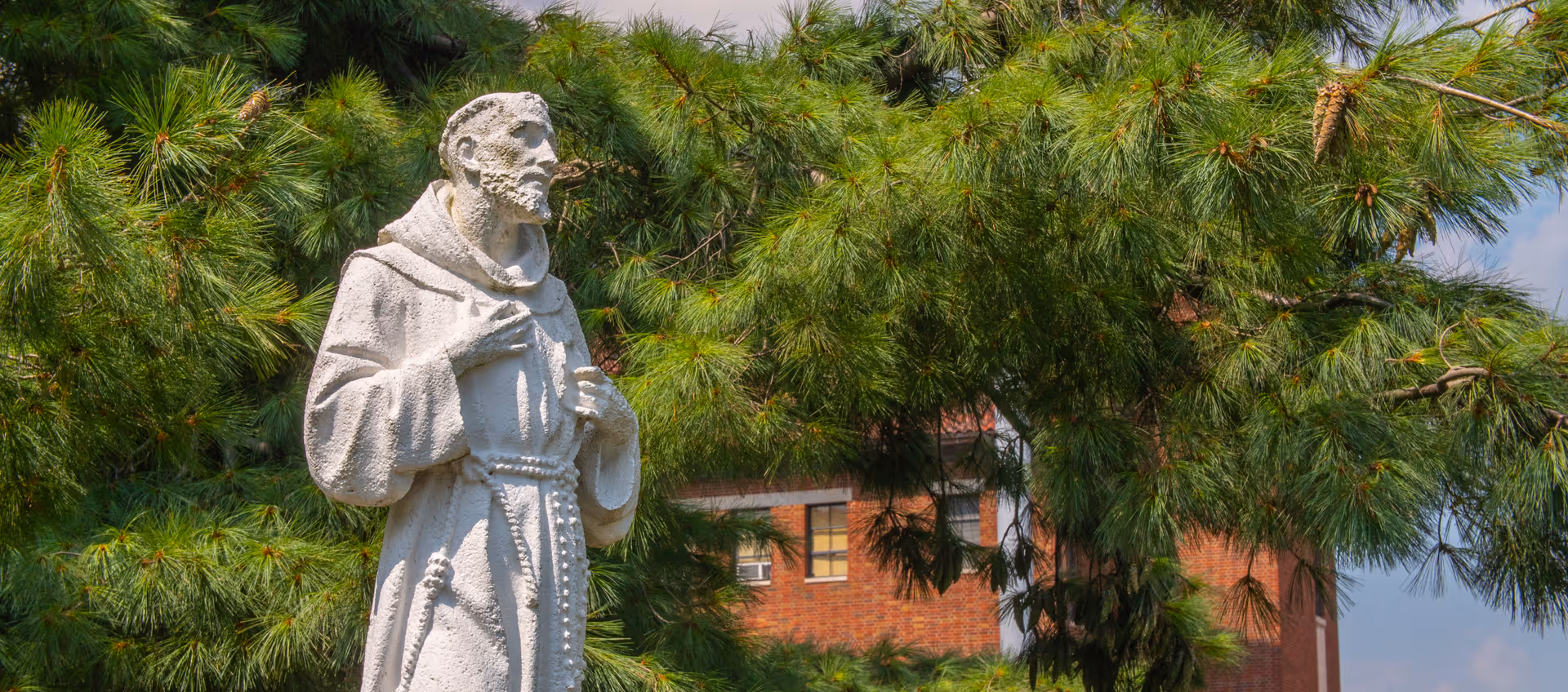 A white stone statue of a robed figure, likely a monk or saint, standing outdoors in front of green pine tree branches with a red brick building partially visible in the background under a blue sky.