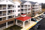 Exterior view of a multi-story senior living facility named Conifer Village at Oakcrest, featuring white siding with stone accents, multiple balconies, a covered entrance with a red roof, and a parking lot with several cars.