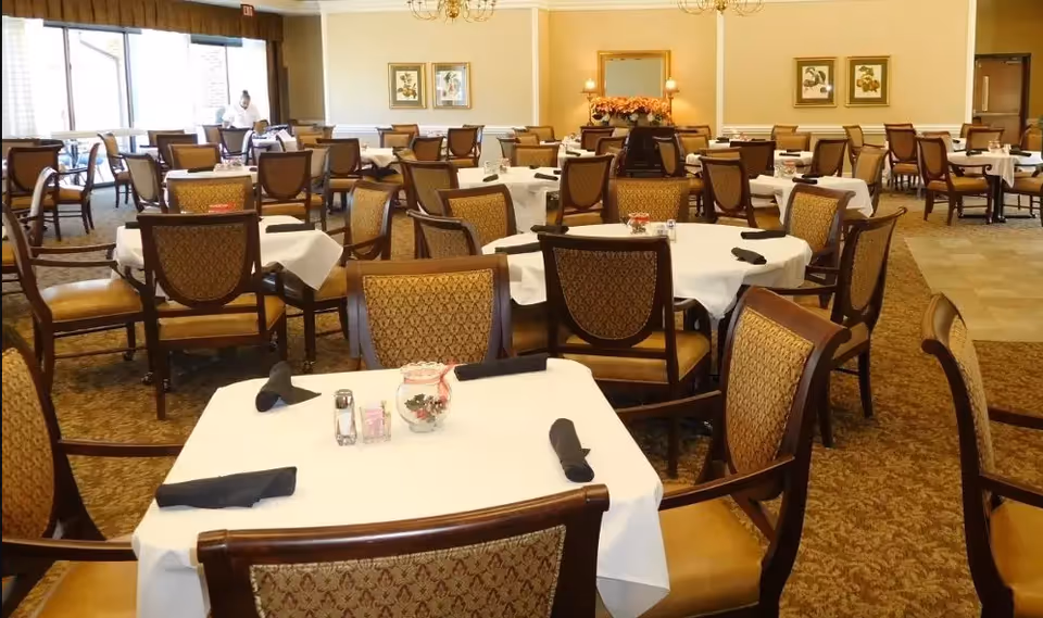 A spacious dining room with multiple round and square tables covered with white tablecloths. Each table is surrounded by wooden chairs with patterned upholstery. The tables are set with black napkins, salt and pepper shakers, and small decorative centerpieces. The room has large windows letting in natural light, framed artwork on the walls, and a carpeted floor.
