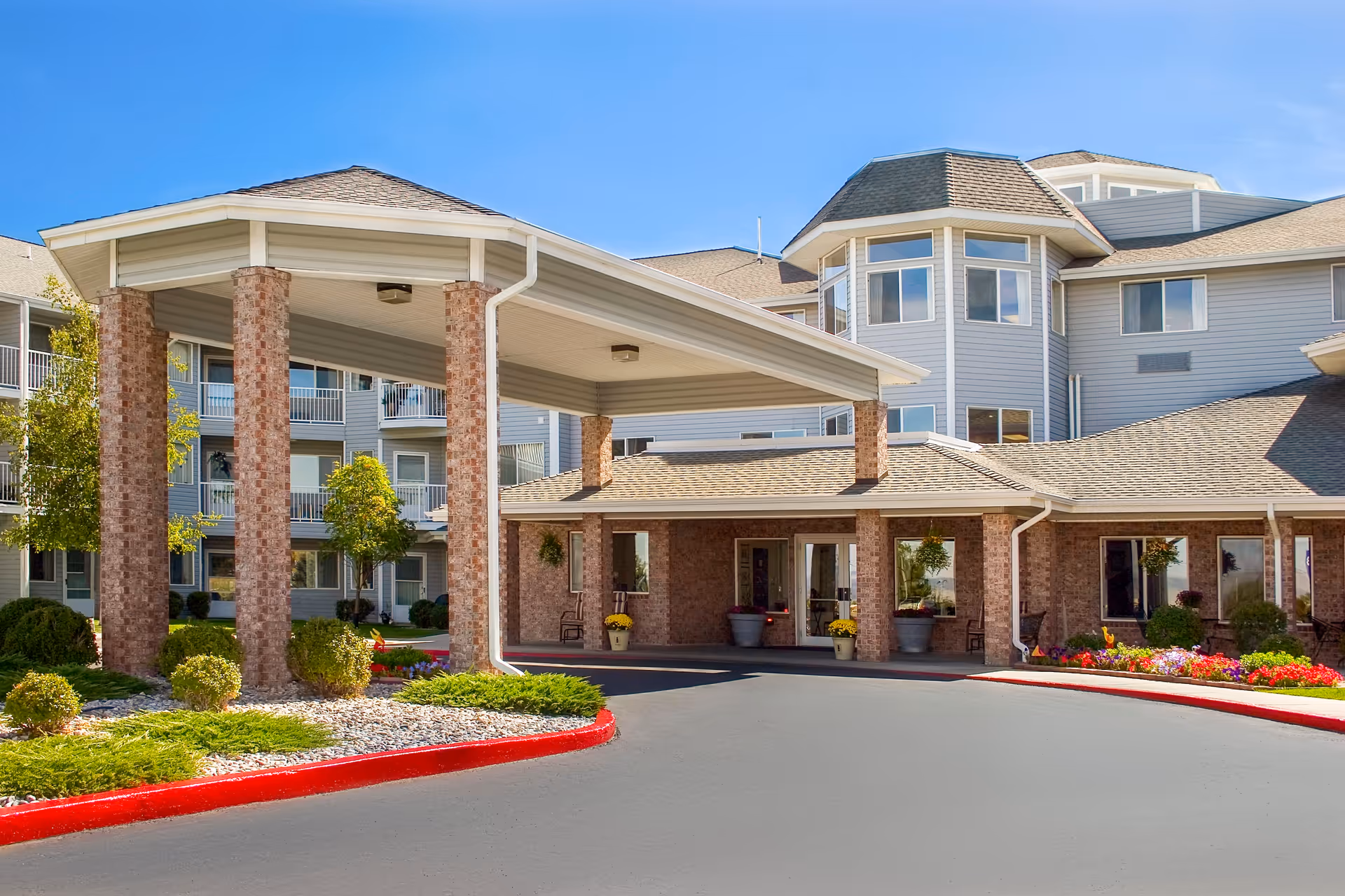 Entrance of Helena Pointe Senior Independent Living facility showing a covered drop-off area supported by brick columns, surrounded by landscaped greenery and colorful flowers, with a multi-story building in the background under a clear blue sky.