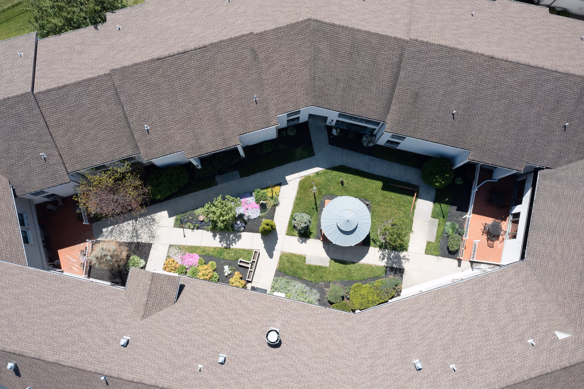 Aerial view of a courtyard garden enclosed by a building with a brown roof. The courtyard features paved walkways, green grass, various shrubs and flowering plants, a central gazebo, and benches. There are also two small patio areas with tables and chairs on either side of the courtyard.