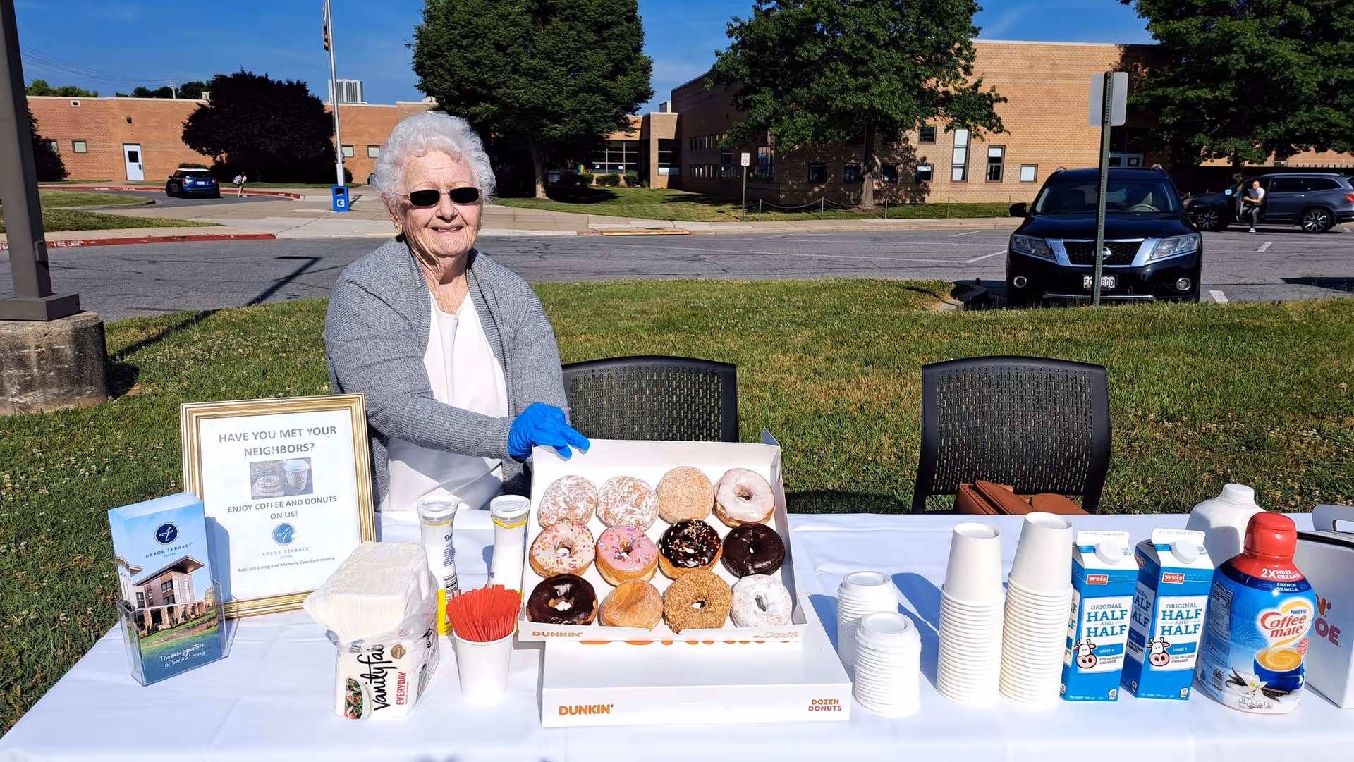 An elderly woman wearing sunglasses and a gray cardigan sits behind a table outdoors with a box of assorted Dunkin' donuts, coffee creamers, cups, napkins, and promotional materials for Arbor Terrace Fulton. A sign on the table invites people to enjoy coffee and donuts to meet their neighbors. The background shows a parking lot, some cars, trees, and a brick building under a clear blue sky.