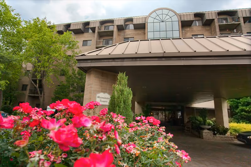 Entrance of Westminster Towers, a multi-story building with a covered driveway. In the foreground, there are vibrant pink flowers and green shrubs. The building has large windows and a distinctive arched window at the top center.