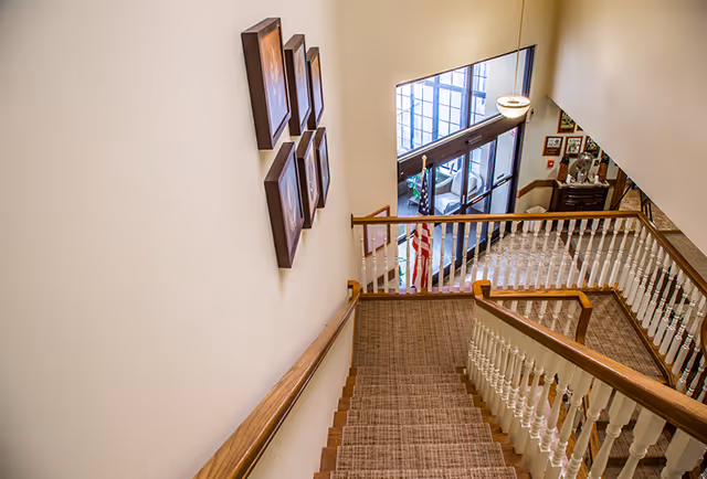 View looking down a carpeted staircase with wooden handrails and white balusters in an assisted living facility. On the left wall, there are six framed pictures arranged in two vertical columns. At the bottom of the stairs, there is a landing area with an American flag near glass doors leading outside. The area is well-lit with natural light coming through the glass doors.
