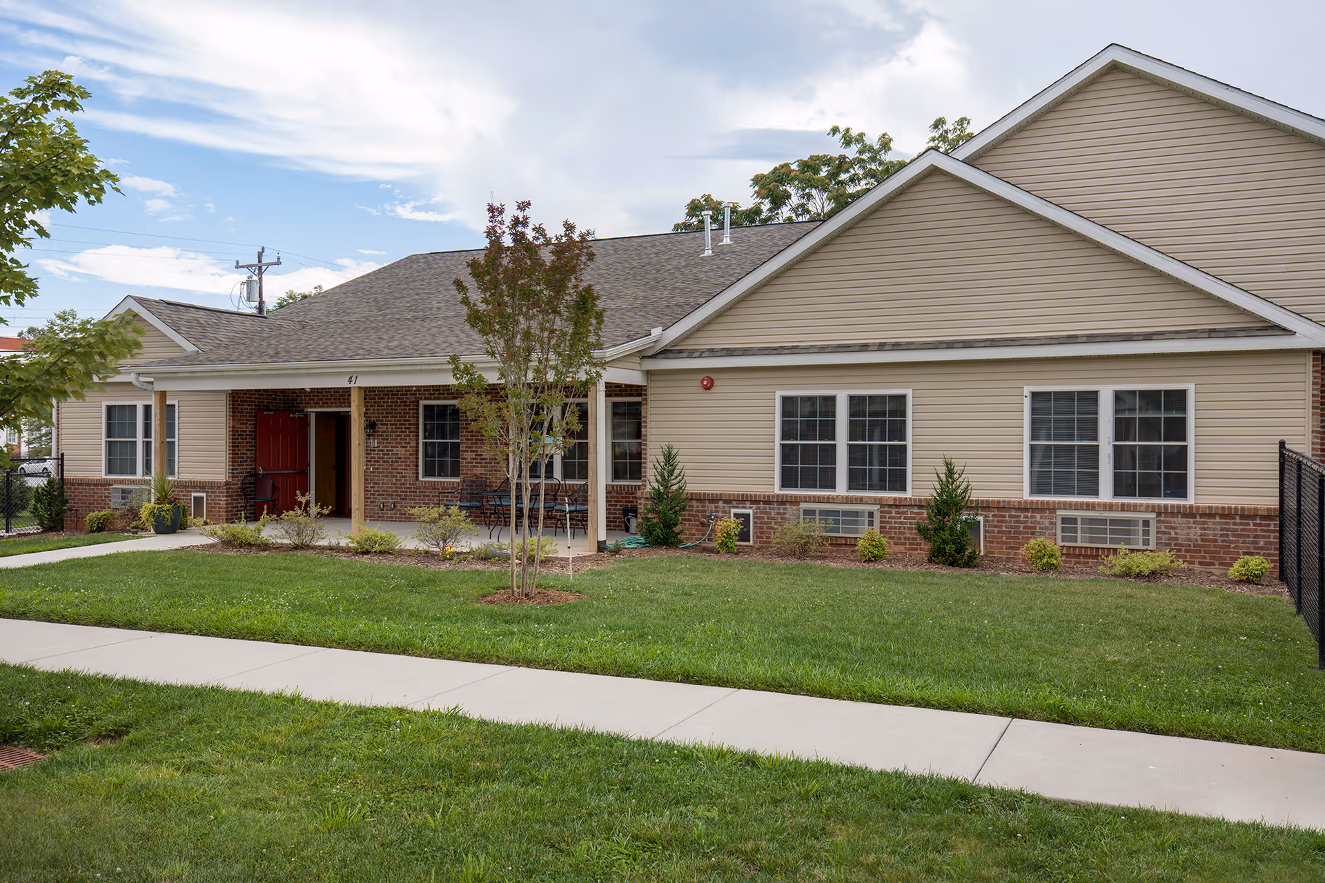 Exterior view of a single-story residential building with beige siding and brick accents, a small covered porch with a red door, several windows, and a well-maintained lawn with a sidewalk in front.