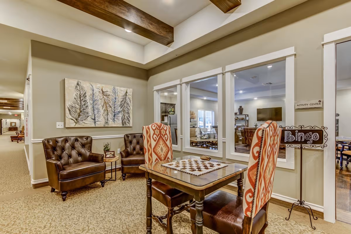 Interior view of a senior living facility seating area with two brown leather armchairs and a wooden table with two patterned upholstered chairs. The table has a checkerboard game set on it. Behind the seating area are large windows looking into an activity center room with a television and more seating. A decorative sign reading 'Bingo' is placed near the windows.