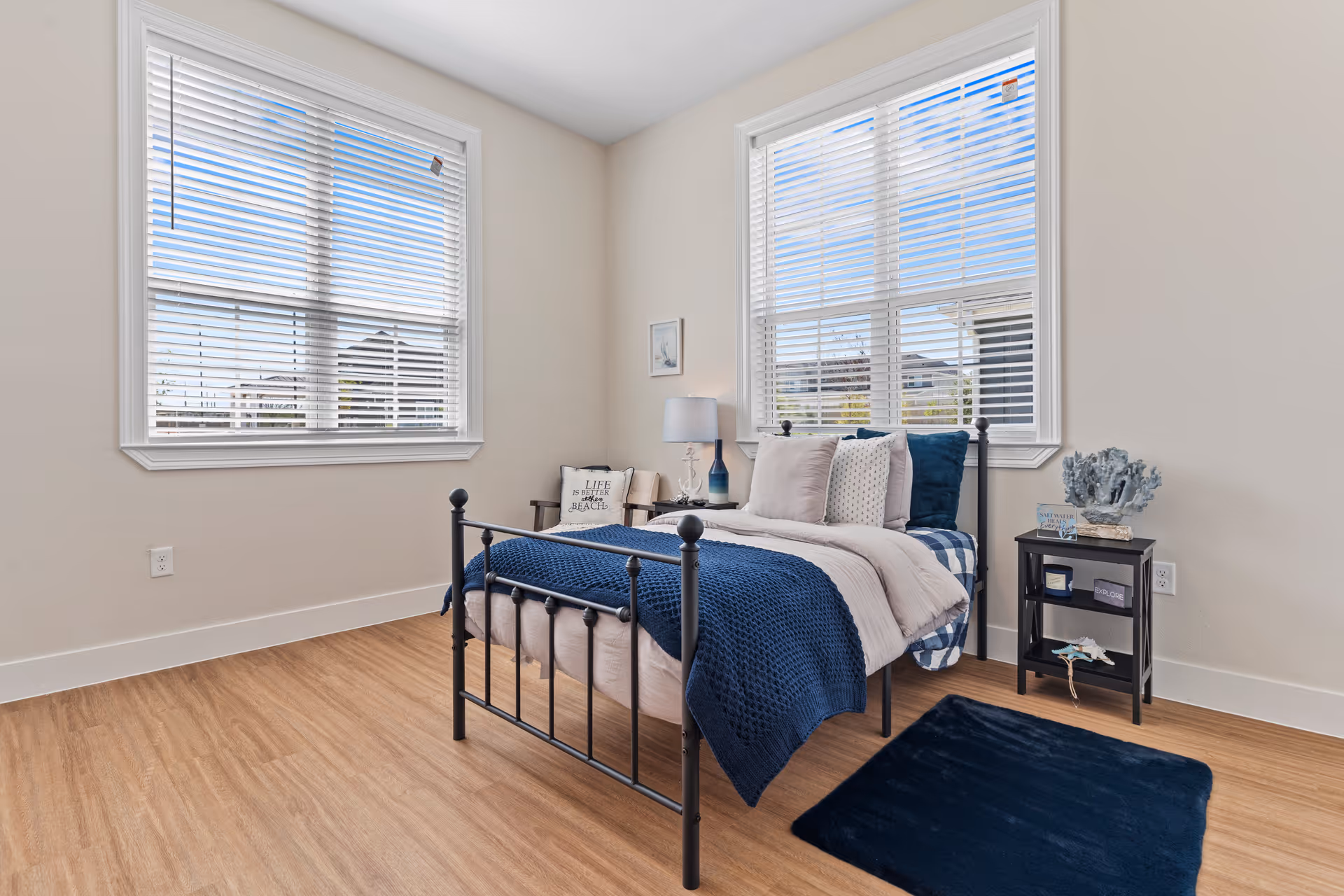A bright bedroom with two large windows with white blinds, a black metal bed frame with beige and navy blue bedding, a small black nightstand with decorative items, a navy blue rug on the wooden floor, and a chair with a cushion in the corner.