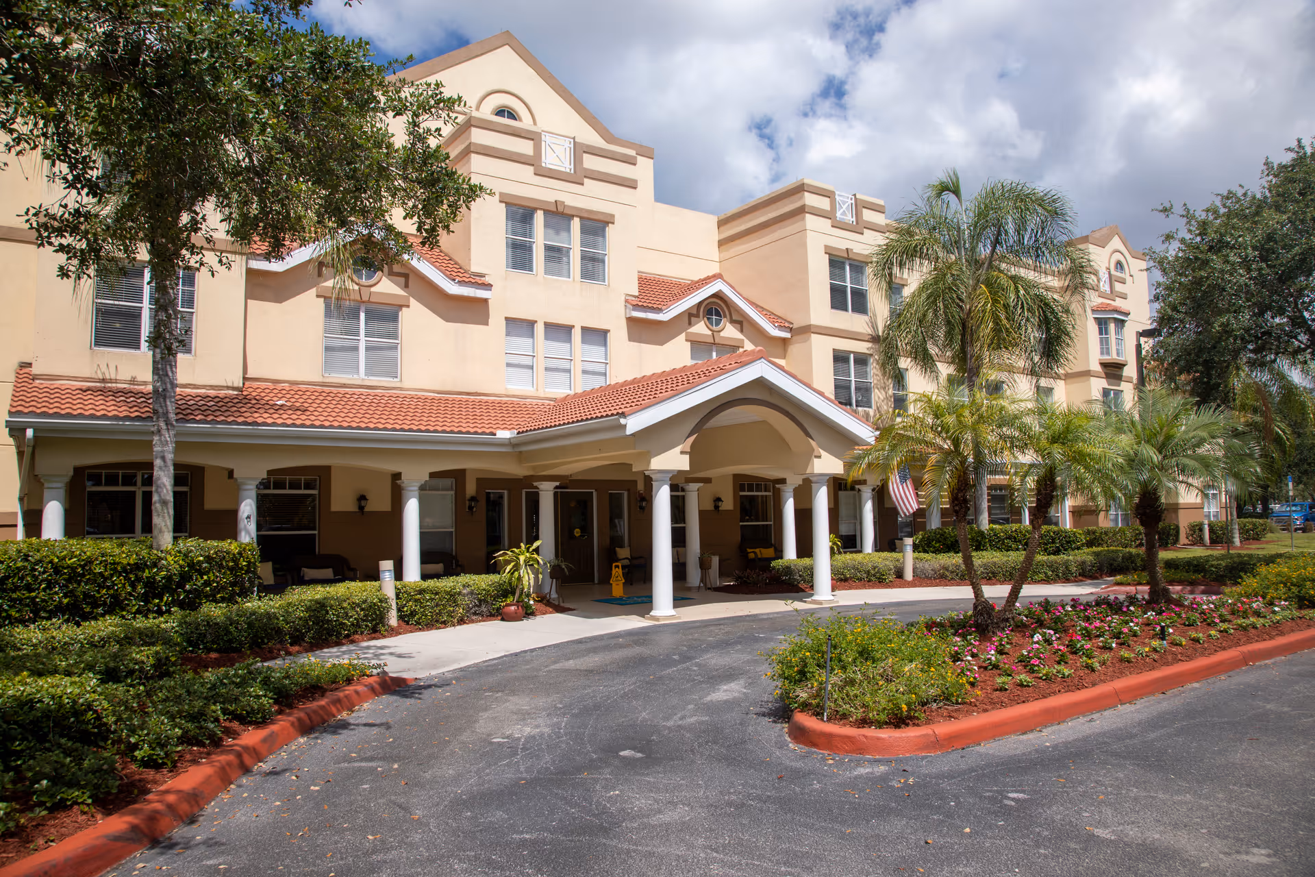 Front entrance of a multi-story beige senior living facility with a covered porte-cochère, palm trees, and landscaped flowerbeds.
