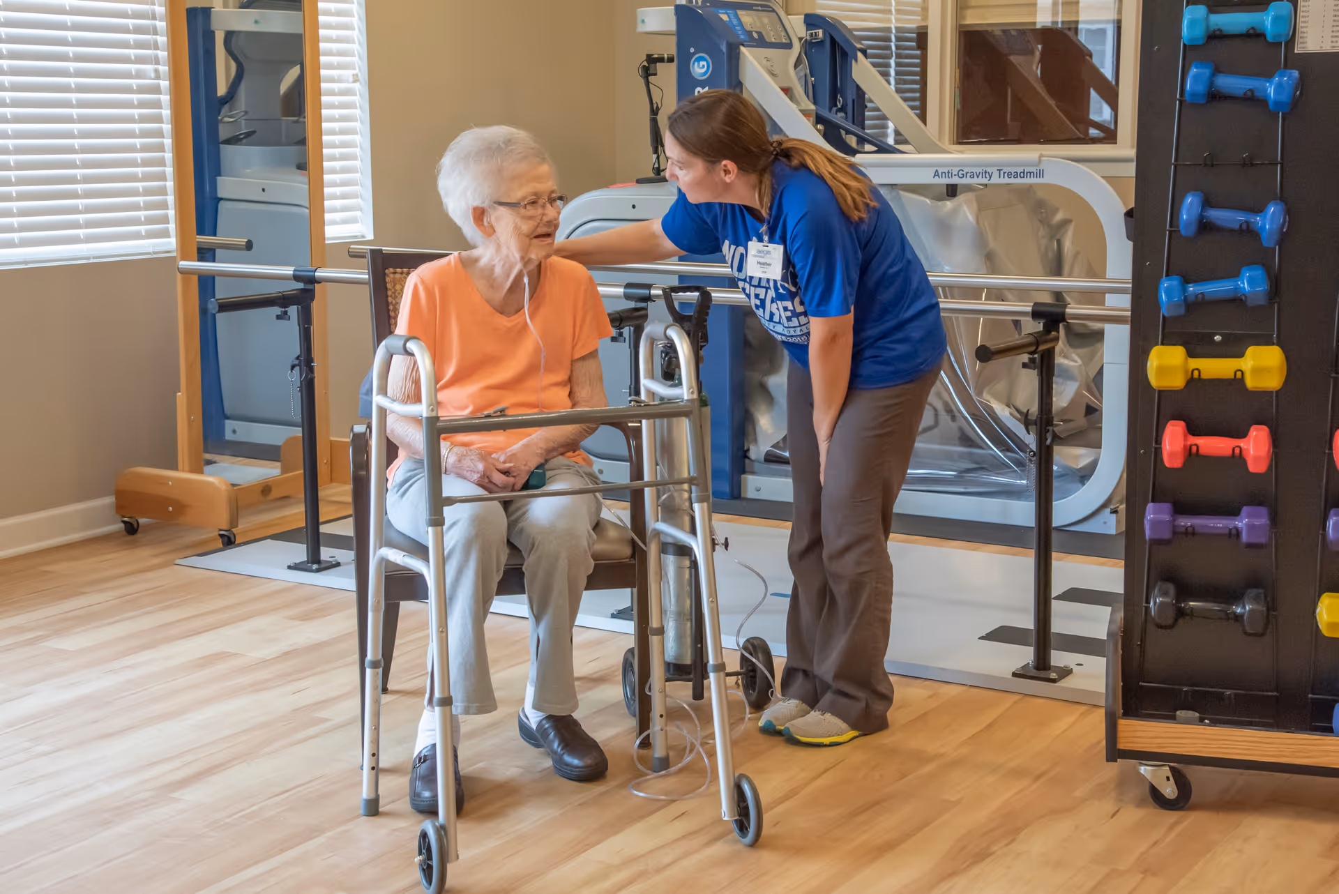 An elderly woman with a walker is seated and wearing an oxygen tube, while a healthcare worker in a blue shirt leans toward her, engaging in conversation in a rehabilitation room with exercise equipment and colorful dumbbells visible.