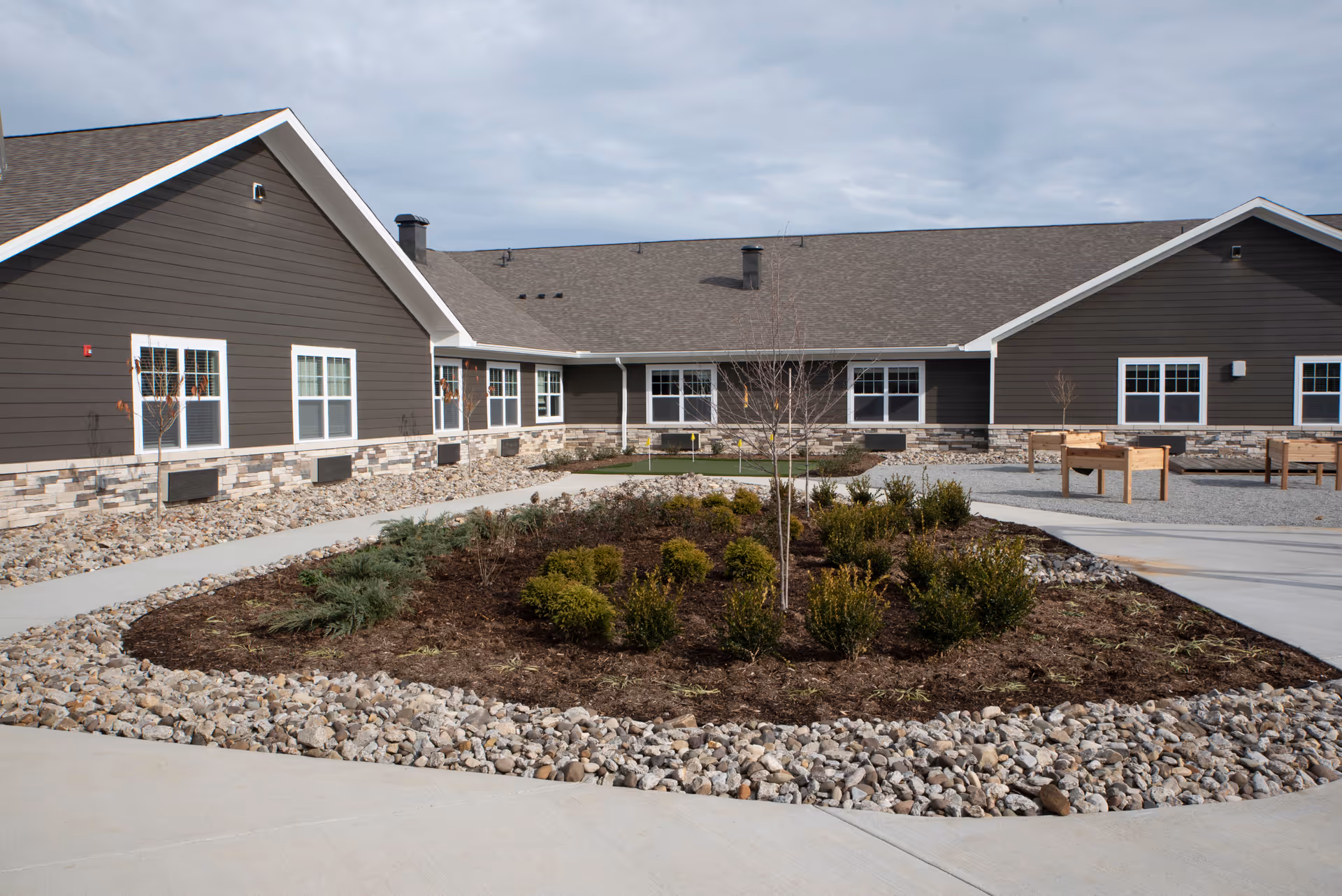 Outdoor courtyard area of a senior living facility with a landscaped garden bed surrounded by rocks and concrete walkways. The building has dark brown siding with stone accents and multiple windows. There are a few small trees and shrubs planted in the garden bed, and raised wooden planters are visible on the right side.