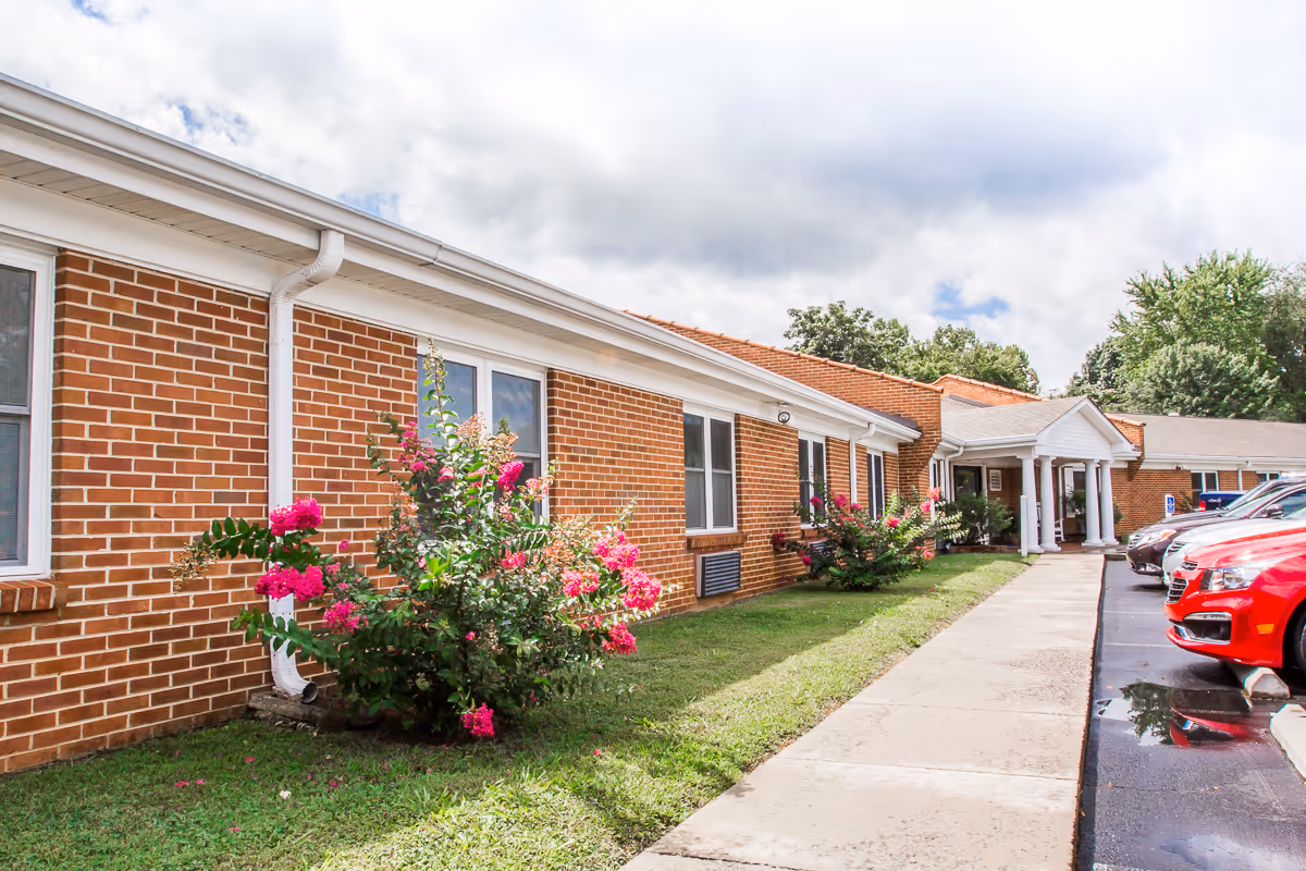Front exterior of a single-story brick senior living facility with flowering shrubs, a sidewalk to a columned entrance, and parked cars.